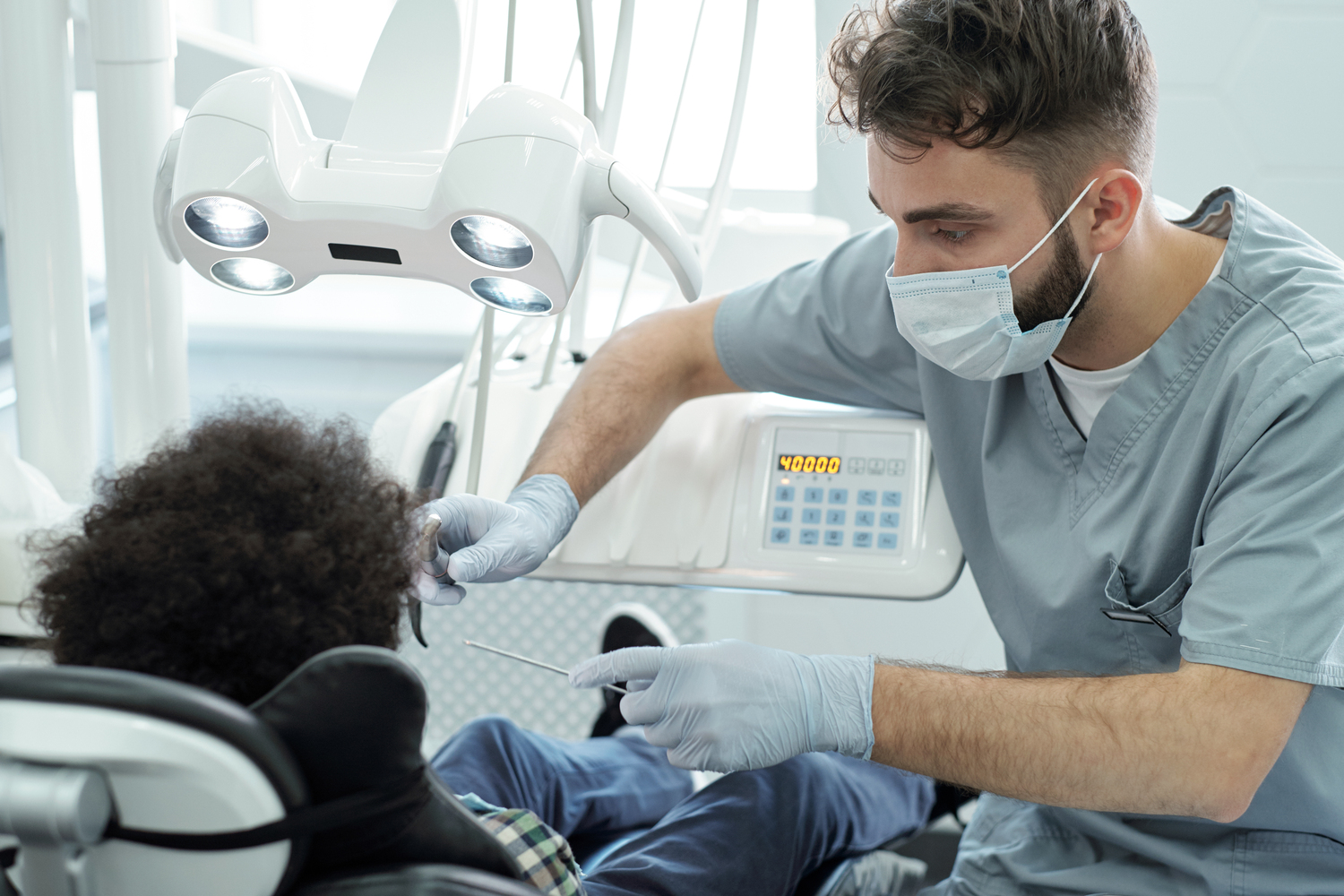 Dentist in uniform, mask and gloves holding dental drill and instrument by mouth of little patient while bending over him