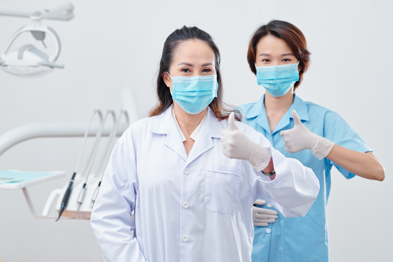 Portrait of cheerful experienced dentist and nurse in protective masks and silicone gloves showing thumbs-up and looking at camera