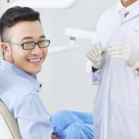 Horizontal shot of cheerful Asian patient of dental clinic sitting on chair smiling, unrecognizable dentist standing next to him