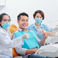 Happy smiling handsome Asian man, his dentist and assistant in medical masks showing thumbs-up after finishing treatment