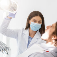 Dentist doctor woman in mask turning on lamp before making check up, patient sitting with open mouth