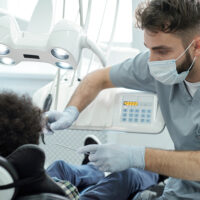 Dentist in uniform, mask and gloves holding dental drill and instrument by mouth of little patient while bending over him